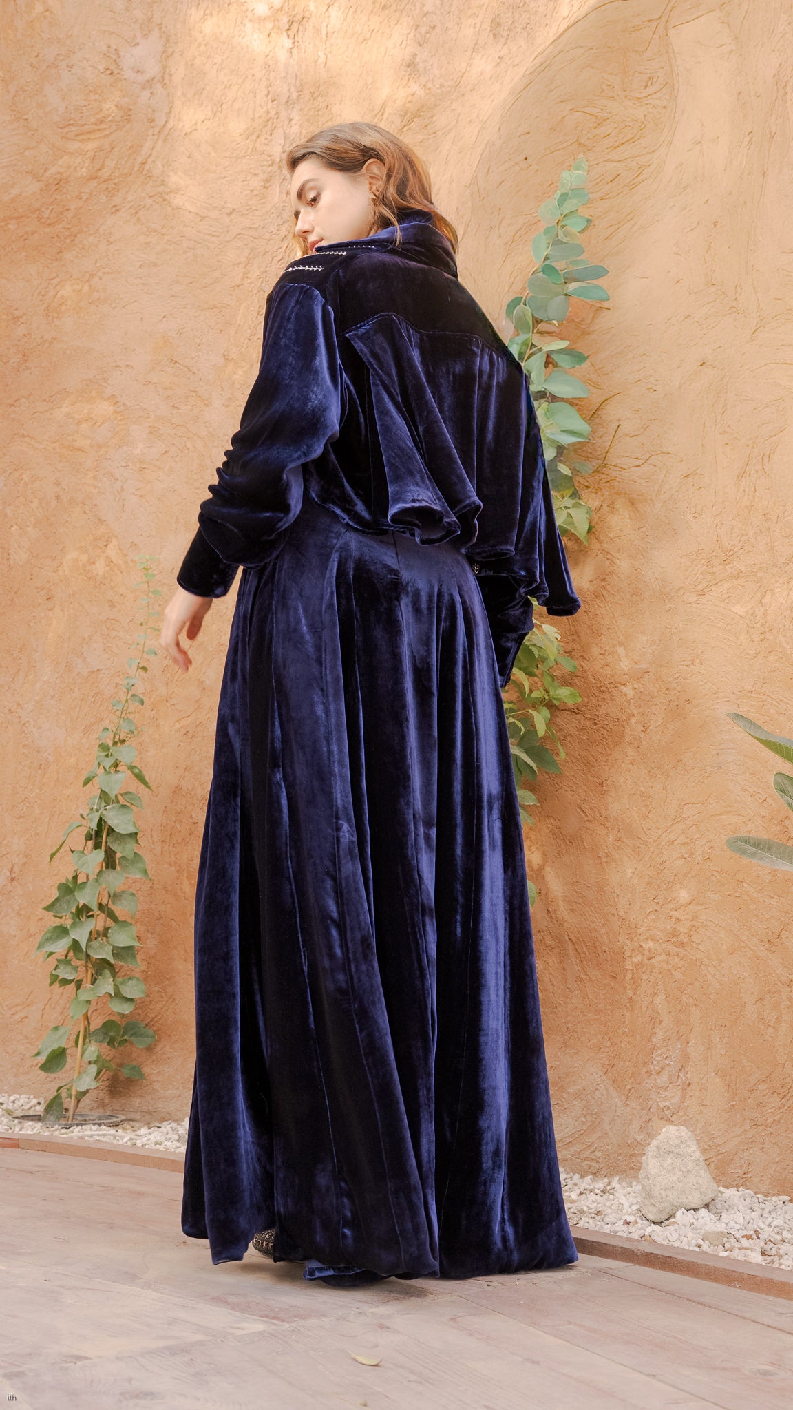 Woman wearing a long navy blue velvet dress against a beige wall with green plants.