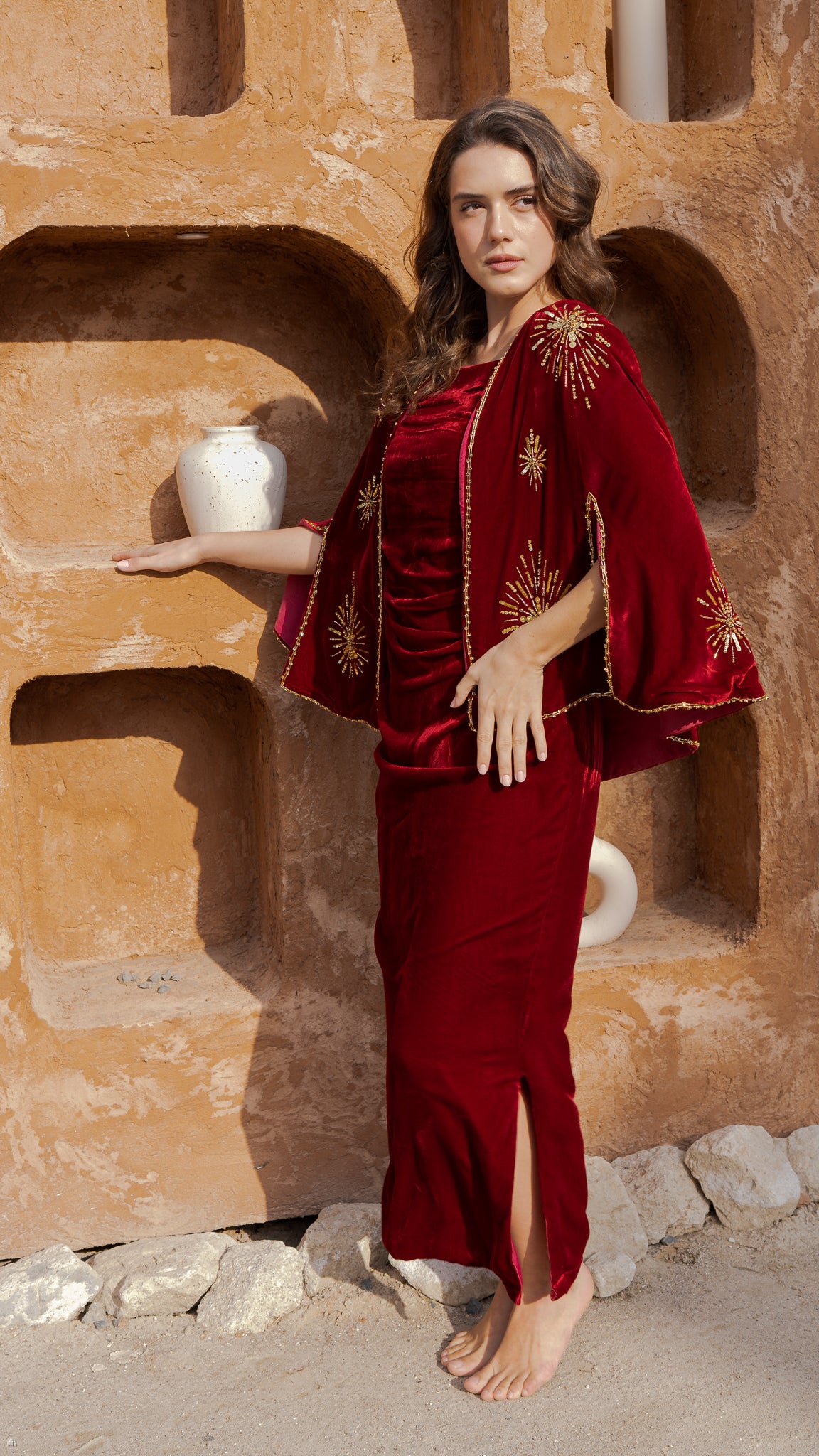 Woman in a red velvet dress with gold embroidery standing against a textured wall.