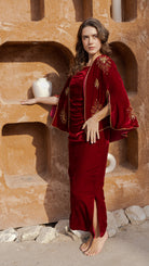 Woman in a red velvet dress with gold embroidery standing against a textured wall.