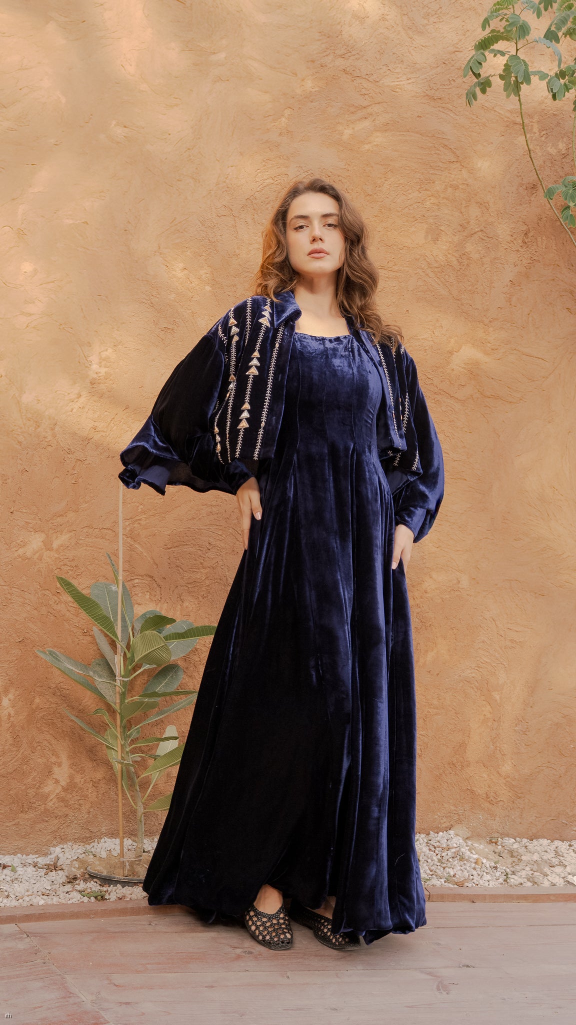 Woman wearing a long blue velvet dress against a beige wall with a plant.