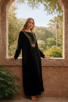 Woman in a black dress with gold embroidery standing in front of an archway with greenery.