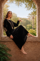 Woman in a black dress with gold embroidery sitting on a stone ledge with a garden view.