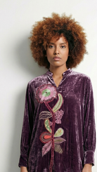 Woman wearing a purple velvet shirt with floral embroidery on a plain background
