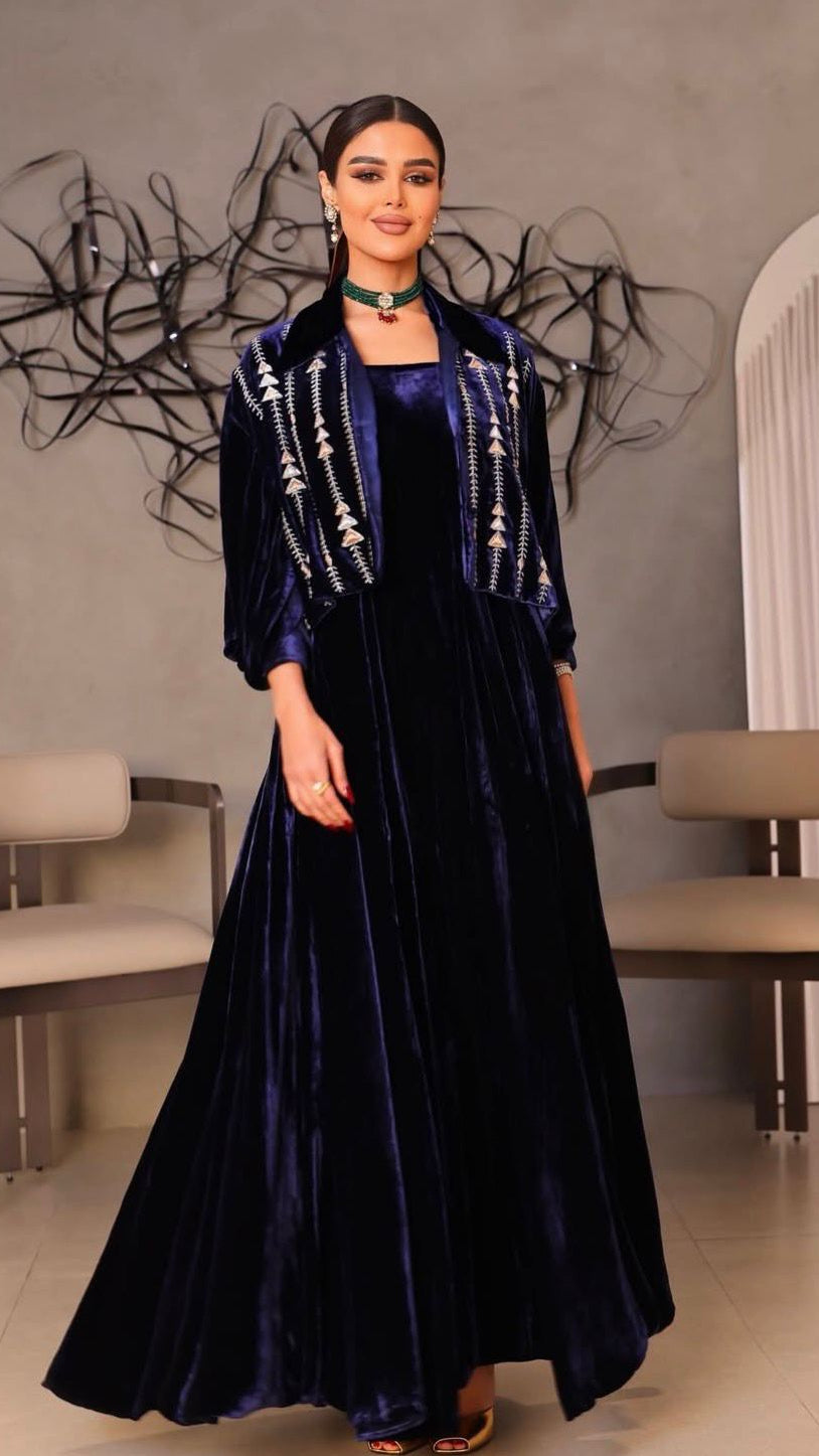 Woman in a dark blue velvet dress with white embellishments standing in a room with chairs and a decorative wall.
