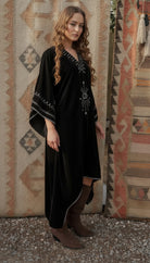 Woman wearing a black dress with intricate designs standing in front of patterned rugs.