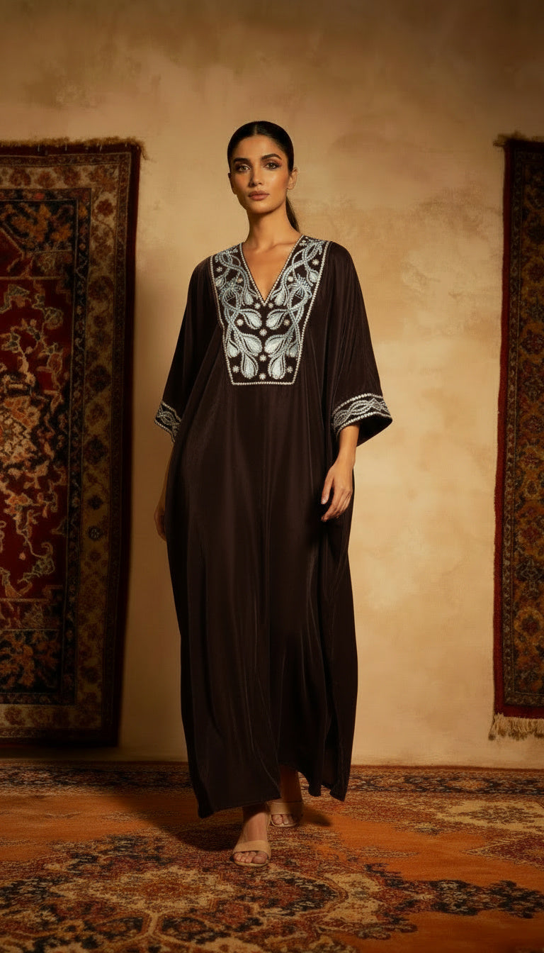Woman wearing a dark brown dress with white embroidery in a room with patterned rugs.